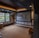 A cozy home theater room features a large projection screen displaying a movie selection interface. The room has dark wood paneling with soft, warm lighting highlighting the ceiling and walls. A large window is situated on one side with an outdoor view partially obscured by trees. There is a curved platform below the screen and a comfortable seating area, including a chevron-patterned ottoman. The room has a neutral color palette with beige, dark gray, and soft brown tones.