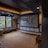 A cozy home theater room features a large projection screen displaying a movie selection interface. The room has dark wood paneling with soft, warm lighting highlighting the ceiling and walls. A large window is situated on one side with an outdoor view partially obscured by trees. There is a curved platform below the screen and a comfortable seating area, including a chevron-patterned ottoman. The room has a neutral color palette with beige, dark gray, and soft brown tones.